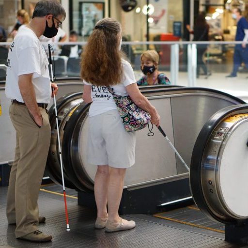 A man teaching a blind woman how to approach an escalator with her cane.