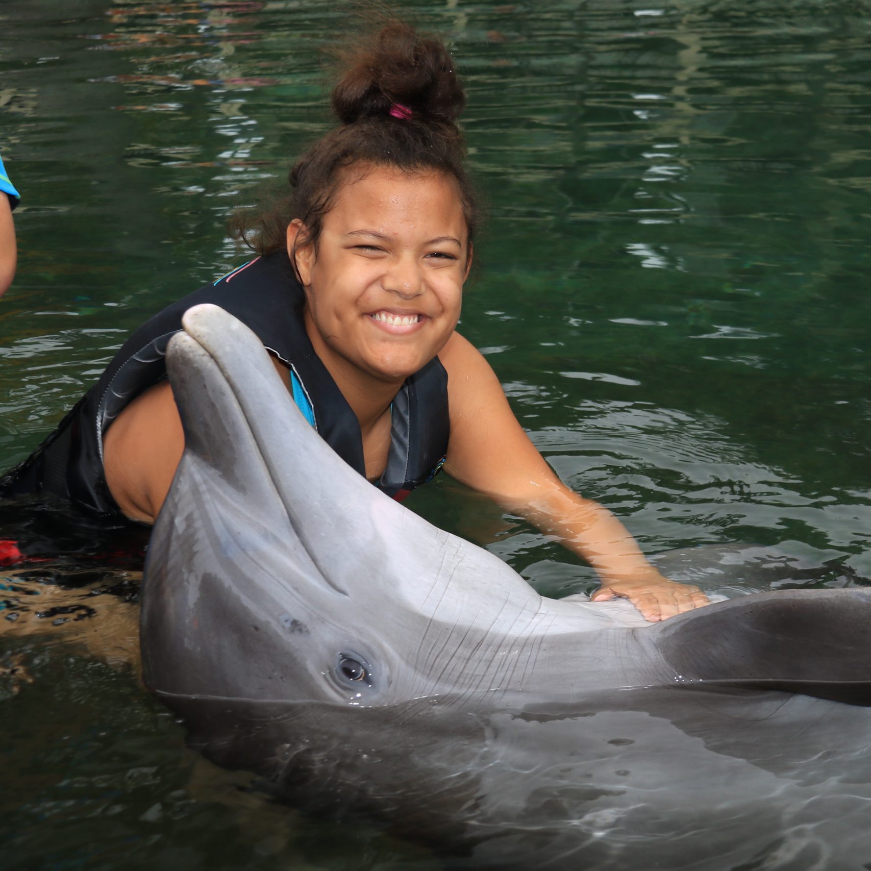 A blind teen girl sits in the water smiling as a dolphin swims next to her.